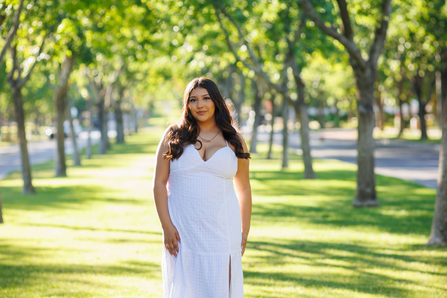 Young woman in white dress poses gracefully in a sunlit green park with tall trees.