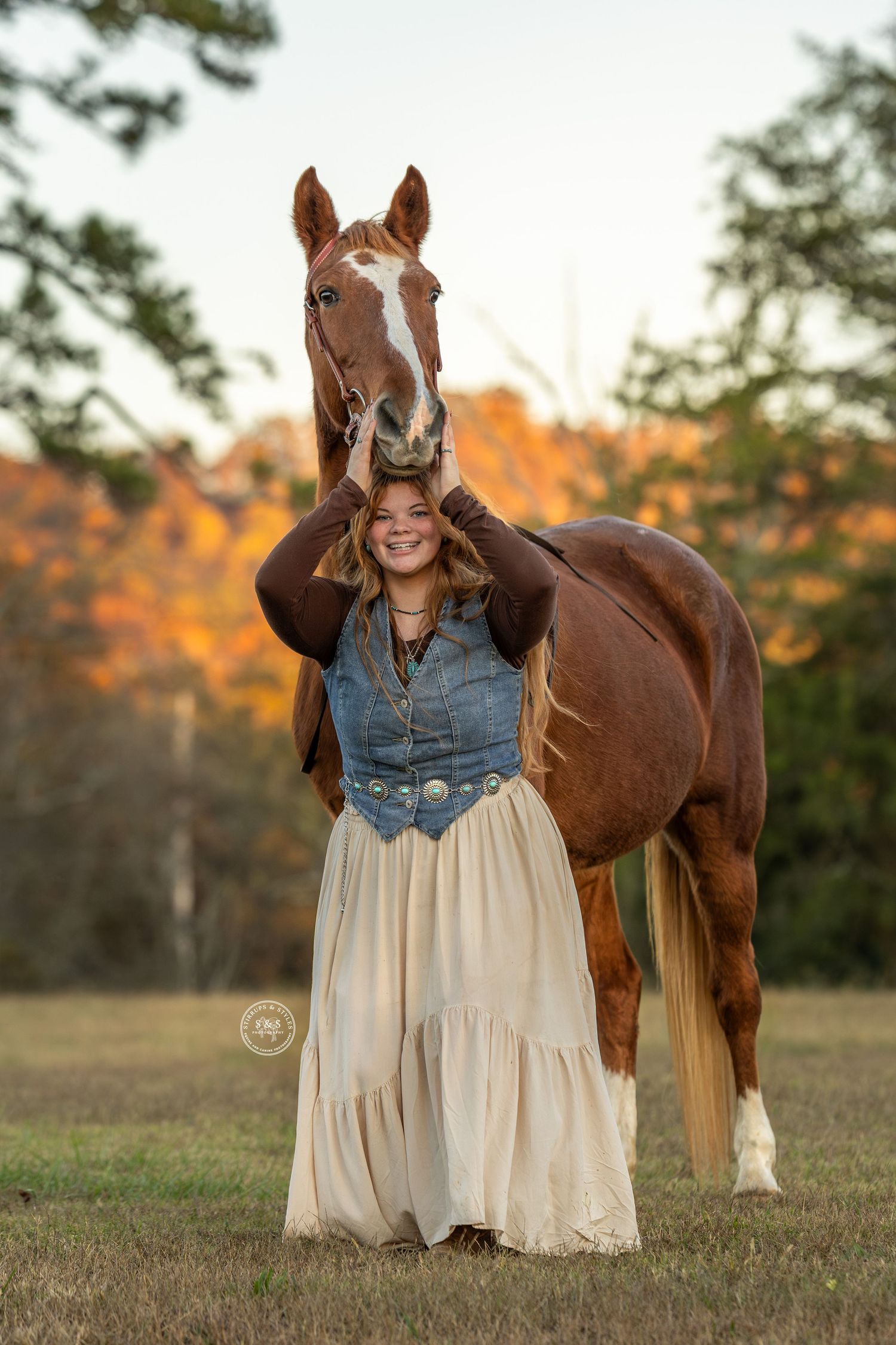 A couple embraces while standing next to a brown horse in a scenic outdoor setting during golden hour.