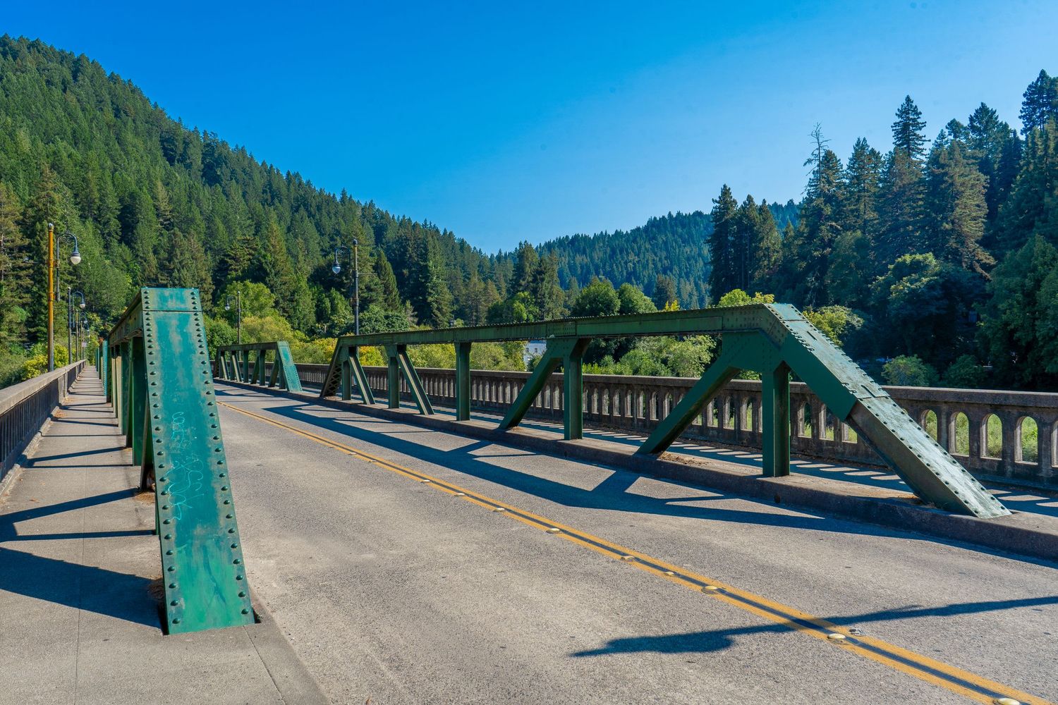 A green steel bridge spans across a river with evergreen forest hills and blue sky in the background. Monte Rio CA