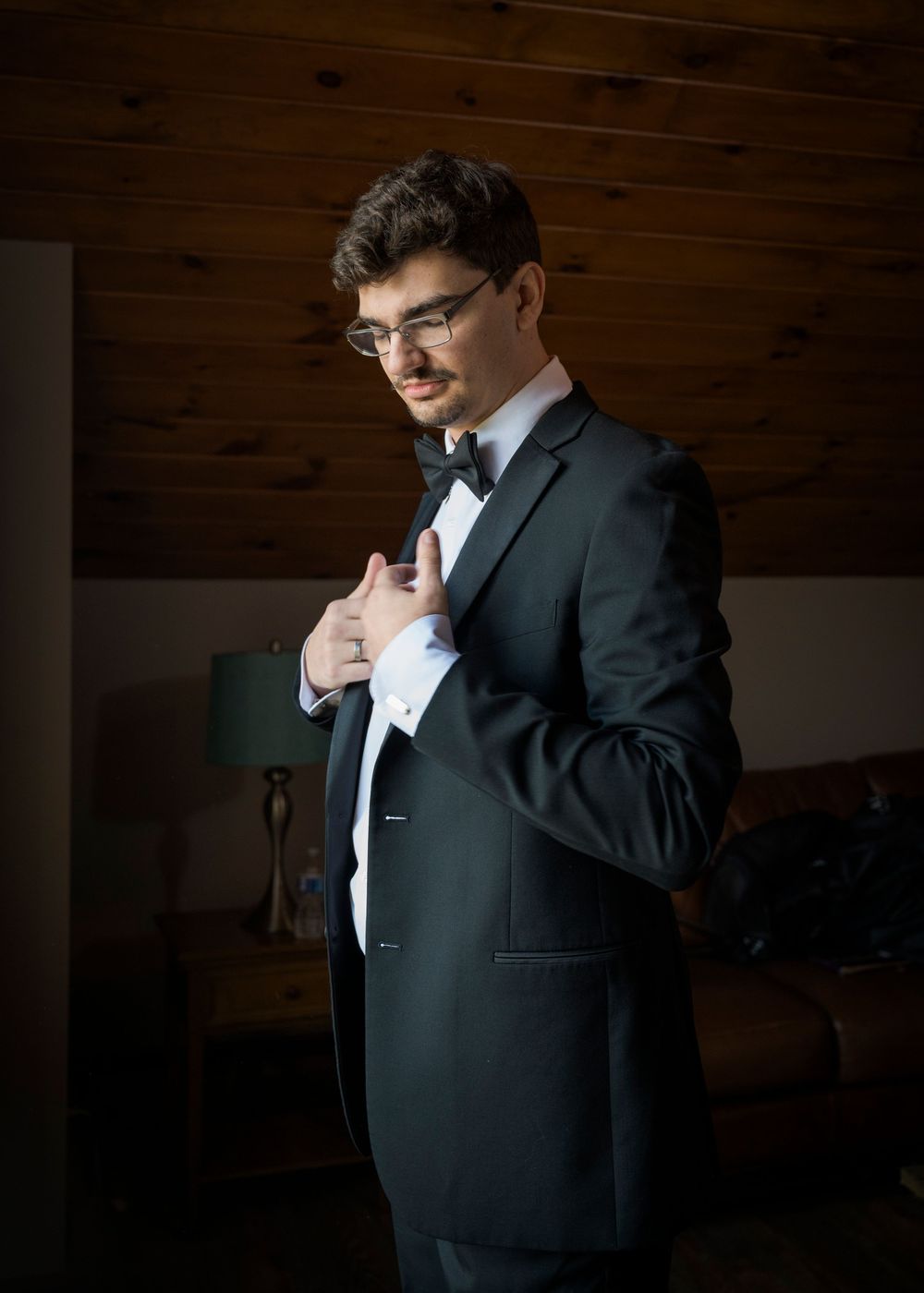 A person in a black suit adjusts their tie while getting ready against a wooden wall background.
