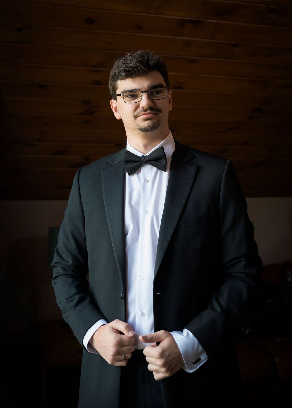 A person in formal attire stands against a dark wood paneled wall while adjusting their clothing.
