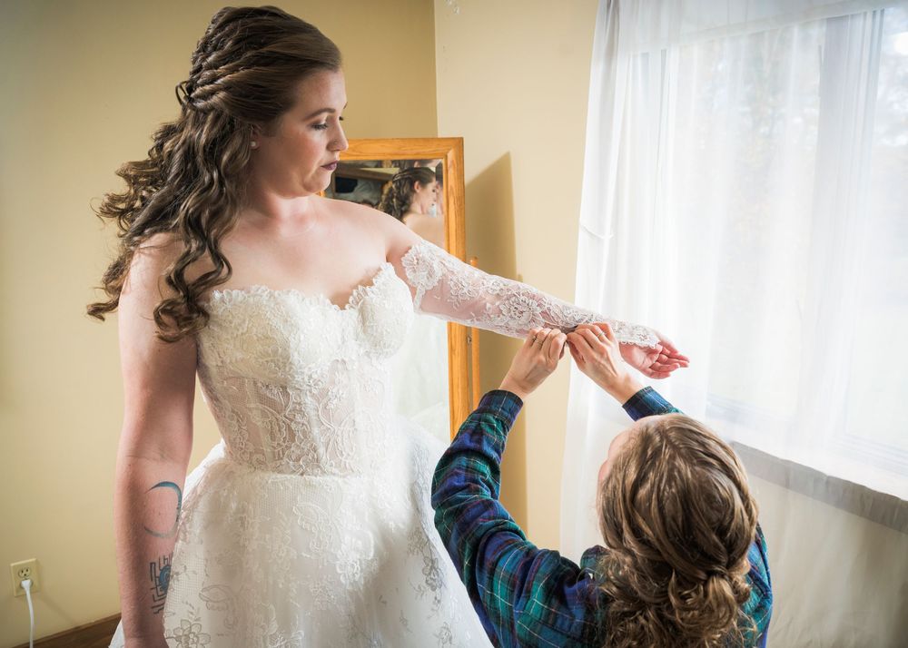 A bride in a white lace wedding dress gets ready in a sunlit room while looking out a window.