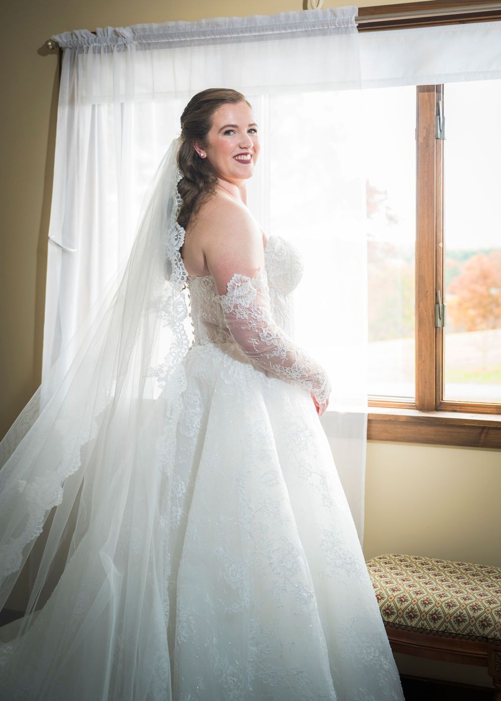 A bride in an elegant white ballgown poses by a window with natural light streaming in.