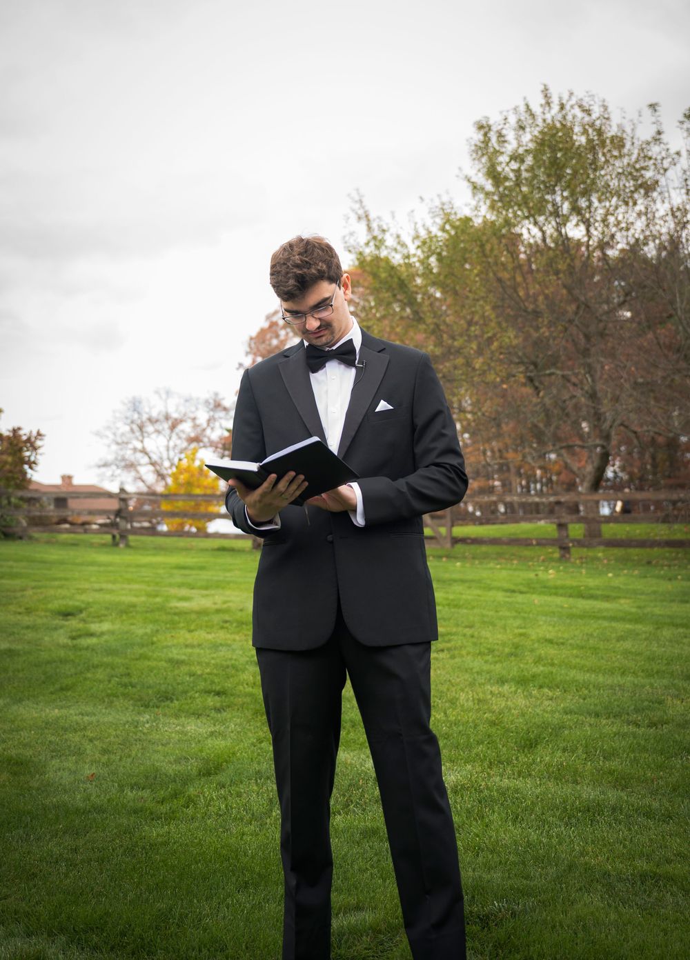 A couple in formal attire reads from books together on a grassy lawn during an outdoor ceremony.