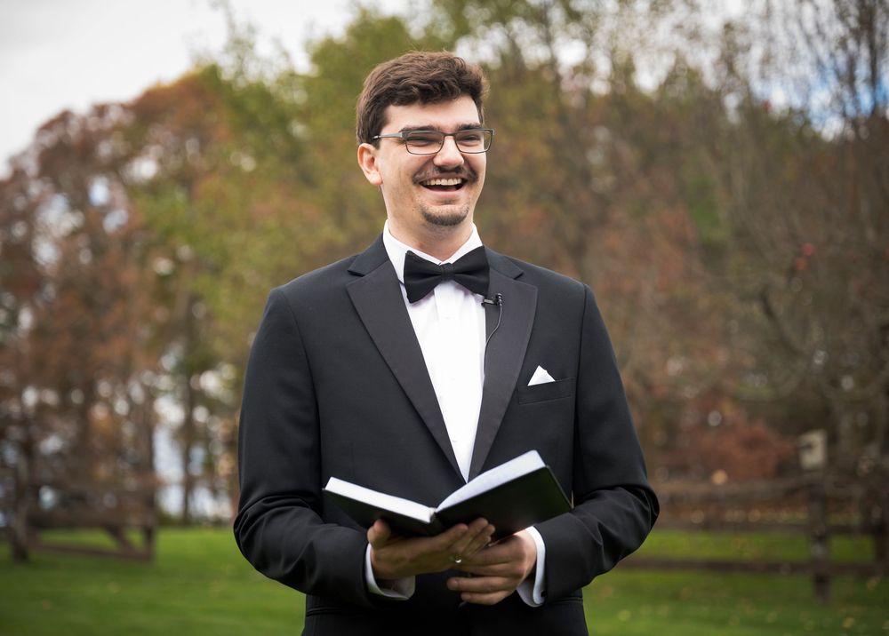 A person in a black tuxedo and bow tie smiles while holding an open book outdoors with autumn trees in the background.