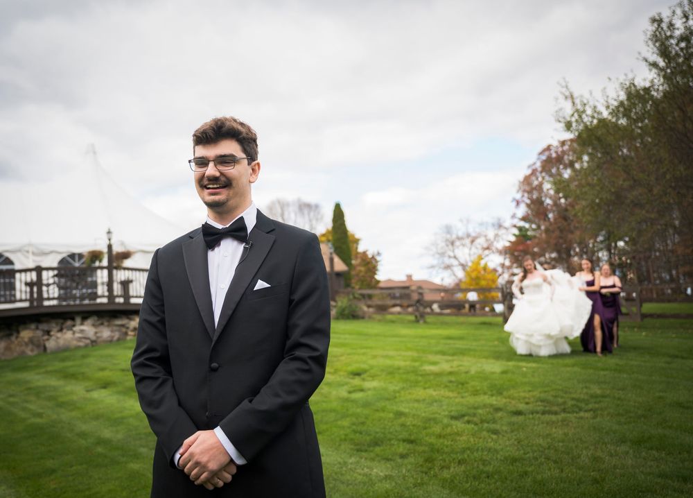 A groom in a black suit poses outdoors on a grassy hill during an autumn wedding celebration.