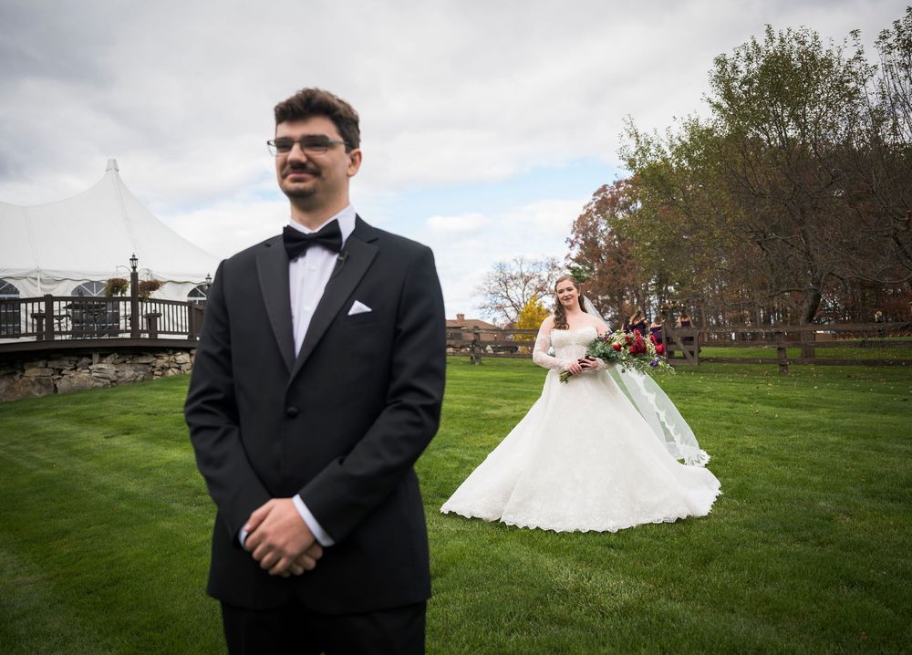 A wedding couple has their first look moment outdoors on a grassy field with autumn trees in the background.