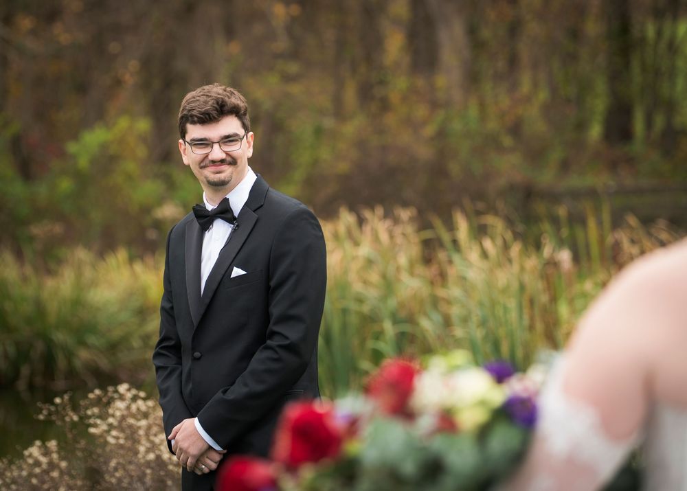 A person in a black tuxedo and bow tie poses outdoors against a natural autumn background.