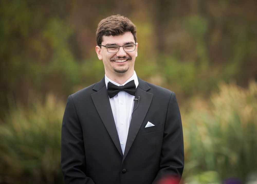 A person in a black tuxedo and bow tie smiles outdoors in front of a natural blurred background.