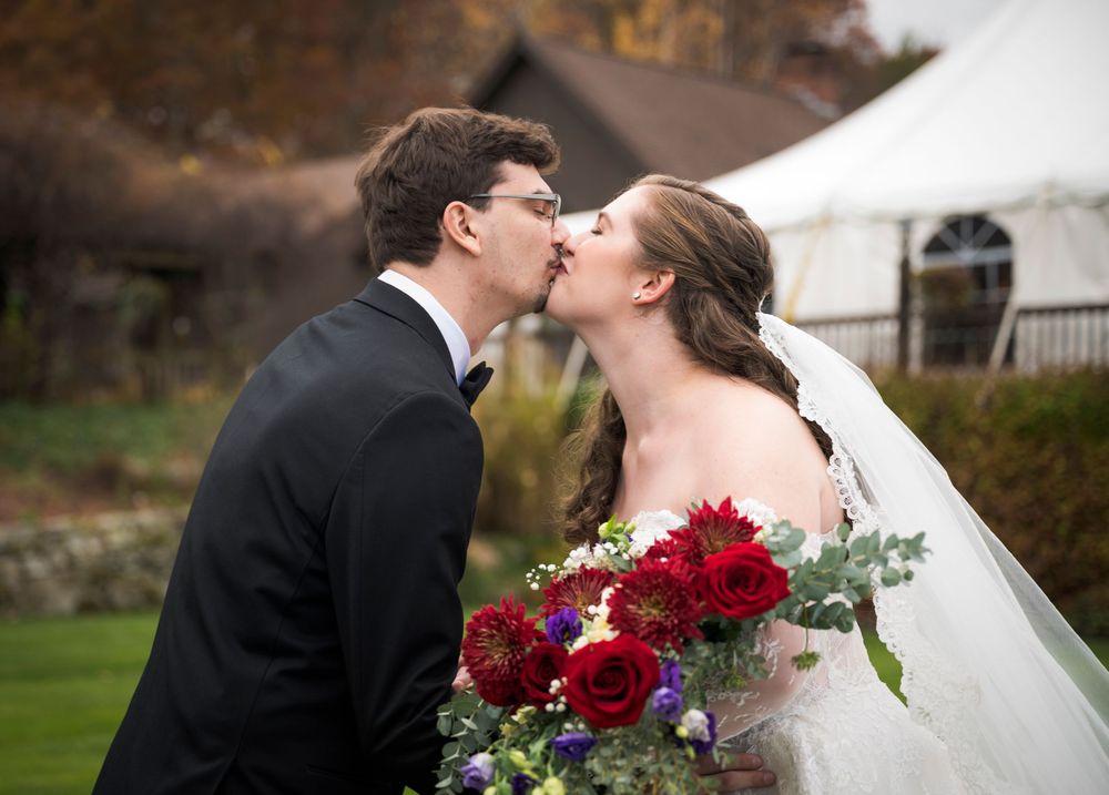 A romantic wedding couple shares tender kisses while holding a vibrant red rose bouquet in an outdoor garden setting.
