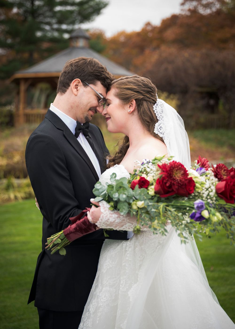 Romantic wedding couple shares an intimate moment outdoors in autumn with red and purple bouquet details.