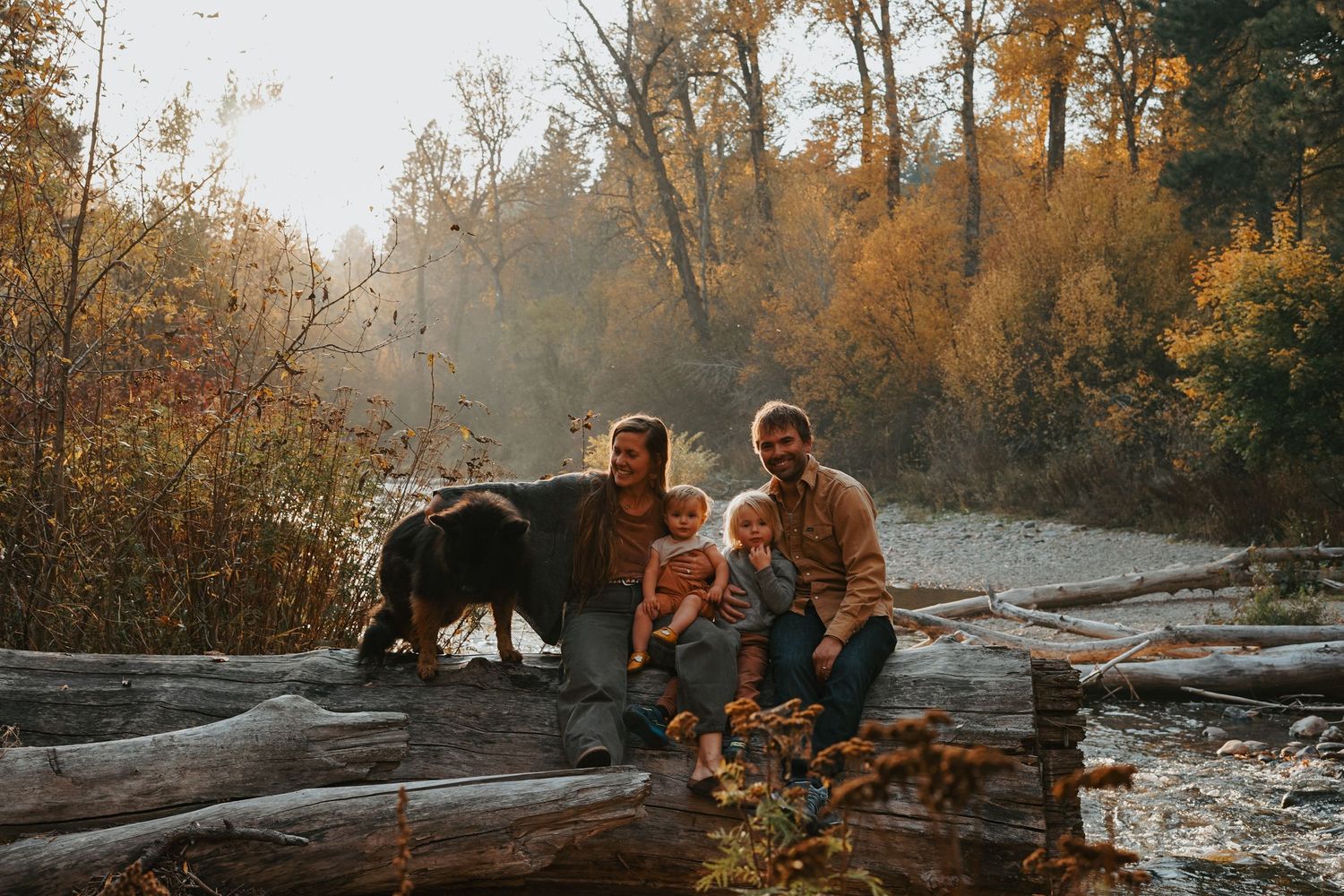 Family sits on fallen logs by a misty river during golden autumn sunset in countryside woods.