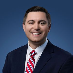 Professional headshot of someone in a navy suit and red striped tie against a blue background.