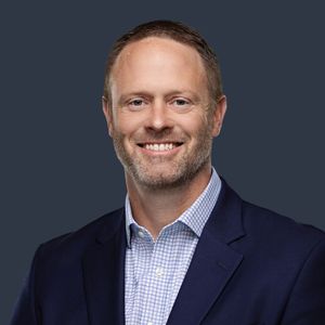 Professional headshot of someone in a navy blazer and checkered button-down shirt with a warm smile.