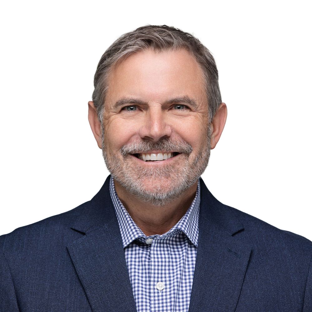 Professional headshot of person in navy suit and blue gingham shirt smiling against white background.