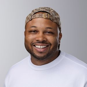 Smiling person wearing a white t-shirt and patterned fabric headband against a gray background.