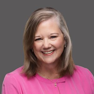 Professional headshot of a woman with gray-brown hair wearing a bright pink top against a gray background smiling warmly.