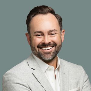 Professional headshot of someone in a light gray blazer and white shirt smiling warmly against a teal background.