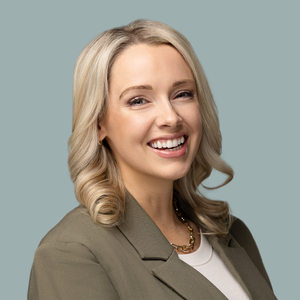 Professional woman in business attire with blonde hair and warm smile poses in a headshot against gray background.