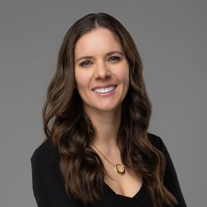 Professional headshot of someone with long dark wavy hair wearing a black top and gold necklace against grey background.