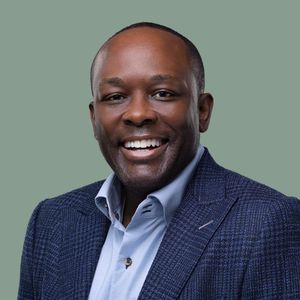 Professional corporate headshot of someone wearing a navy blazer and light blue shirt against a sage green background.