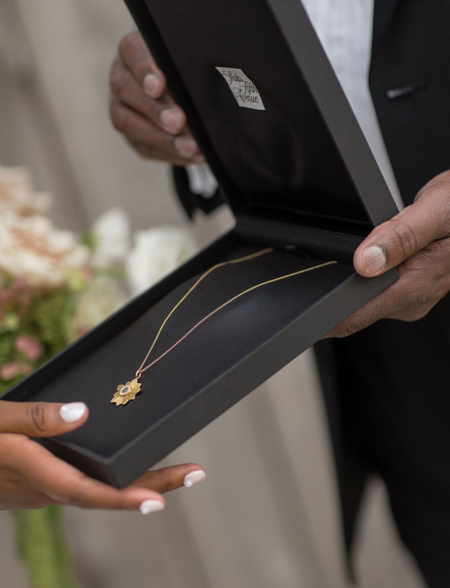 Hands holding open a black jewelry box displaying a delicate gold necklace with pendant against blurred floral background.