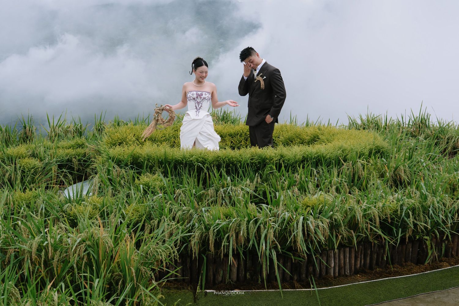 Bride and groom walking through lush green rice terraces with misty mountains in background.