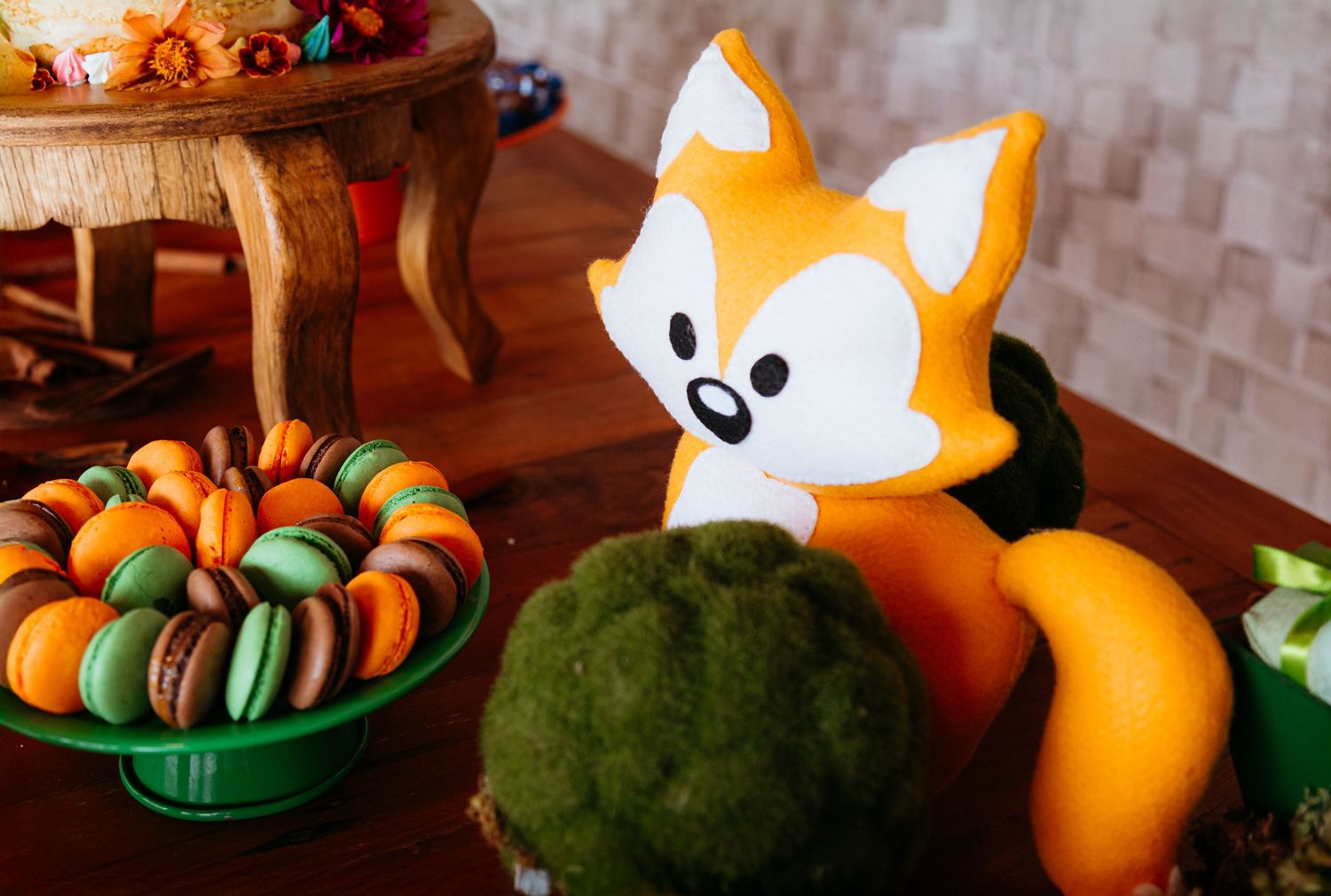 Sweet dessert party display features colorful macarons in green bowl and decorative orange fox figure with fruits.