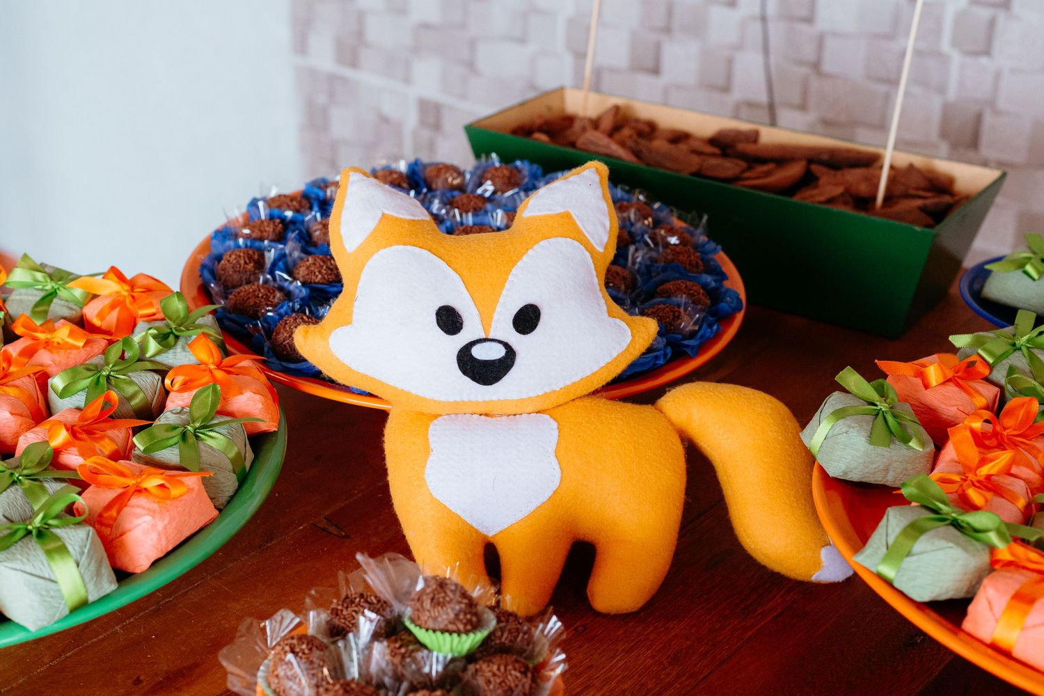 A cute orange plush fox toy sits among colorful fruits and vegetables on a brown wooden table.