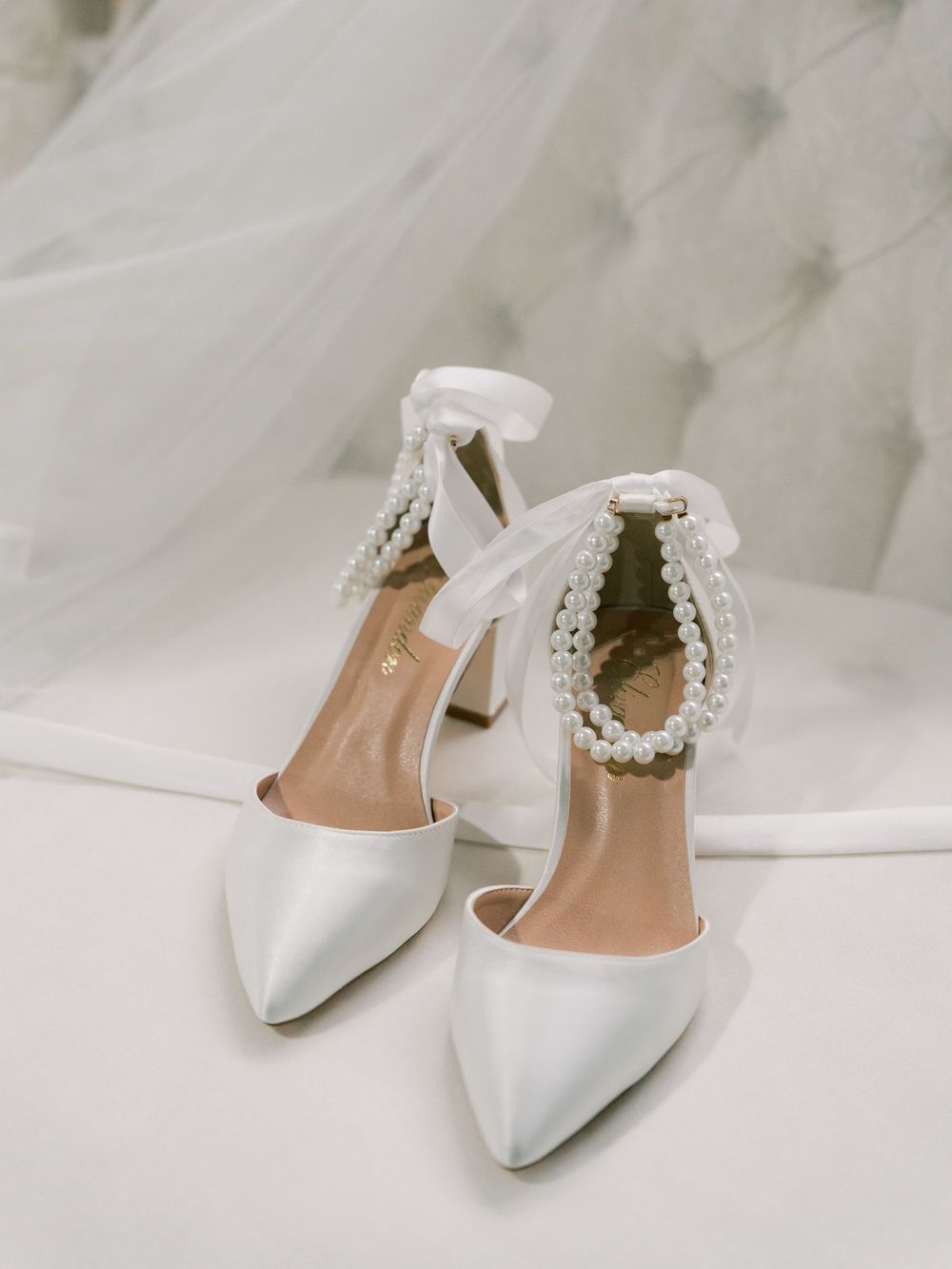 Elegant white bridal shoes with pearl ankle straps on light background.