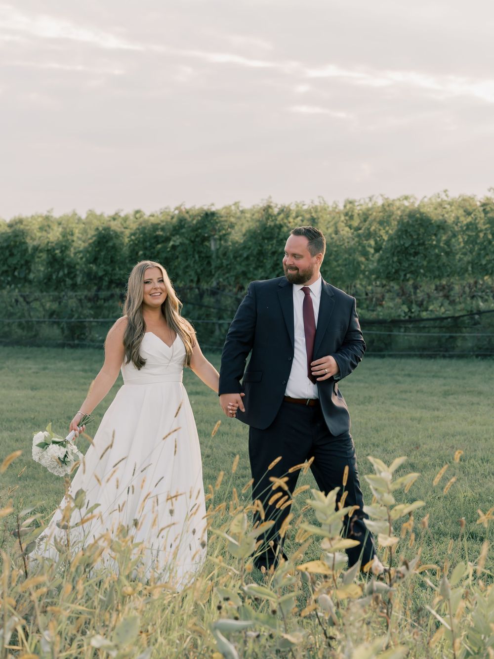 A couple runs playfully through tall grass in a field during their wedding photoshoot.