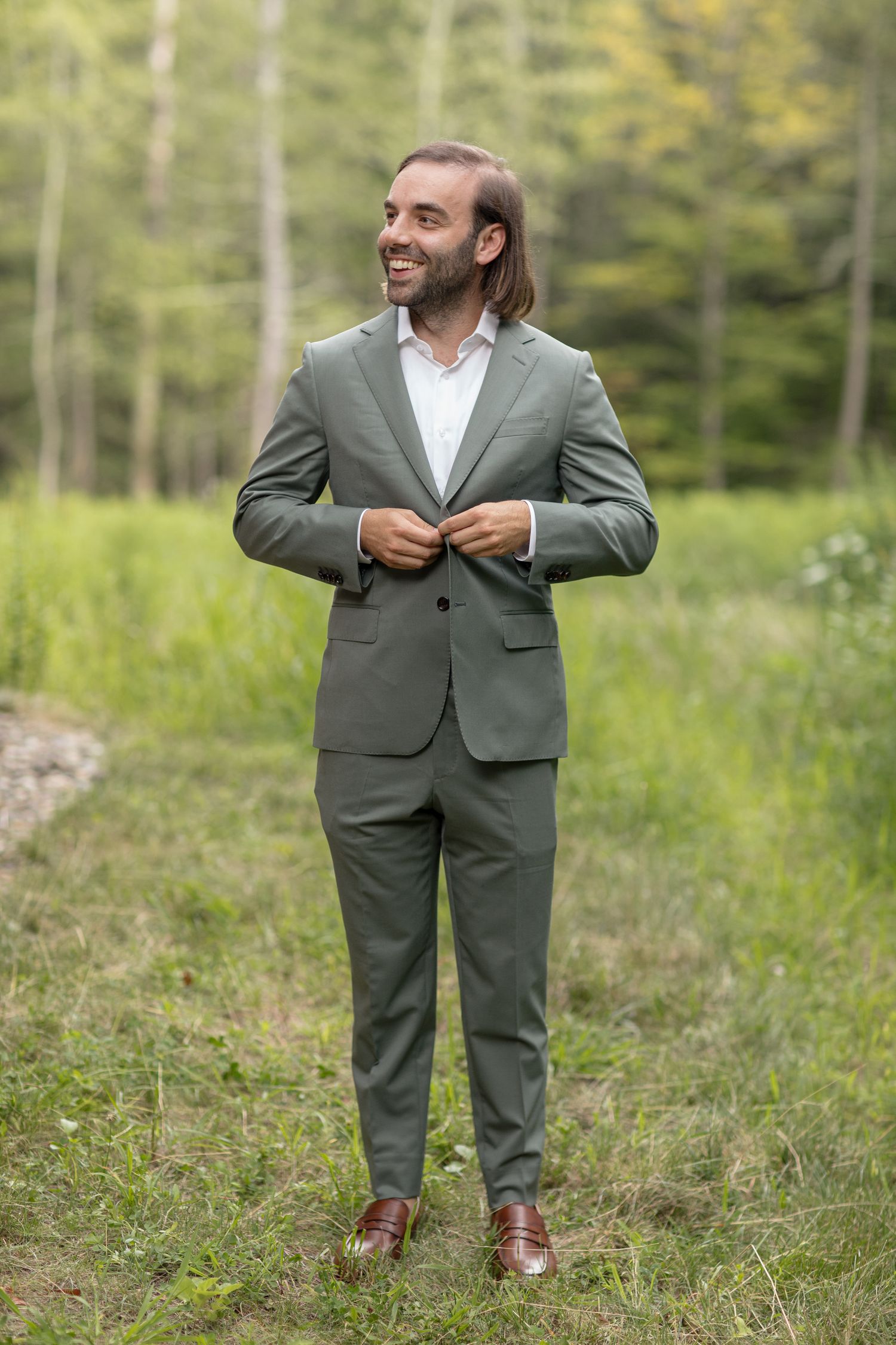 A person wearing an olive green suit stands on a grassy hillside during a casual outdoor photo session.