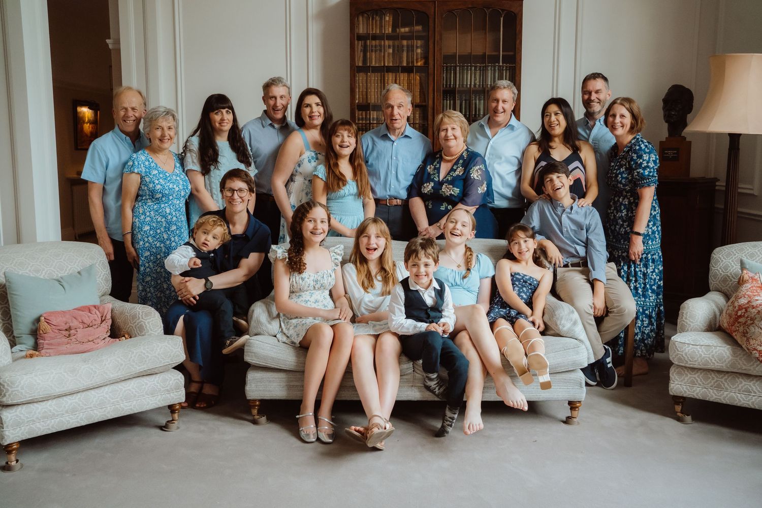 Large group photo showing multiple people sitting together on couches and the floor in a bright living room setting.