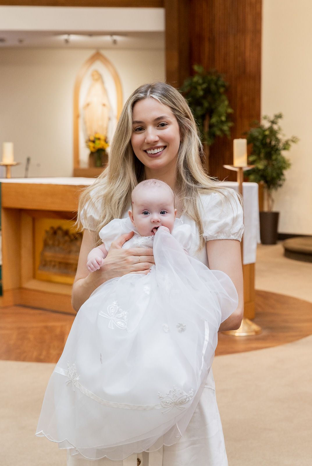 A baby in an elegant white christening gown is held in a church sanctuary setting.