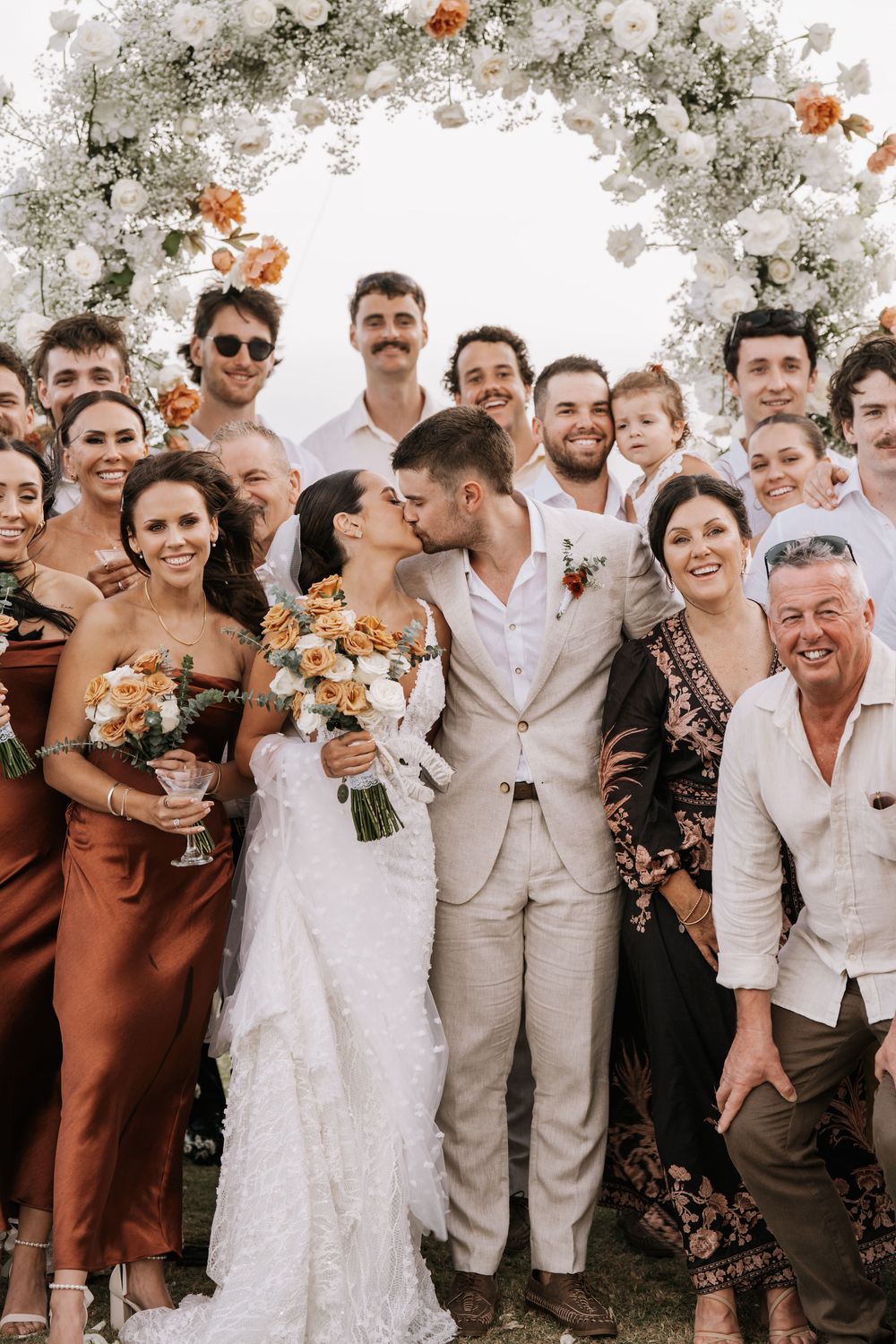 Wedding party gathers together outdoors under white floral arrangements for a festive group photo celebration.