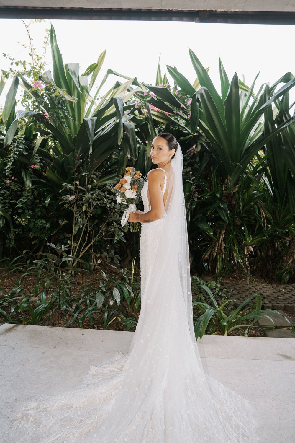 A bride in a white wedding gown with long veil poses in front of tropical greenery and plants at multiple angles.