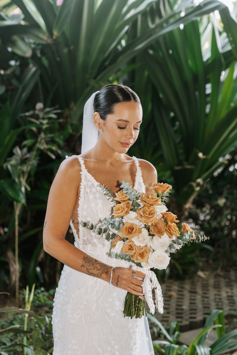 Elegant bride in lace dress holds a bouquet of peach roses against tropical greenery backdrop.