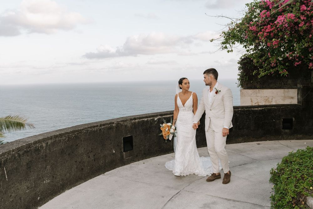 Wedding couple stands by stone wall overlooking tropical beach at sunset with red flowers in background.