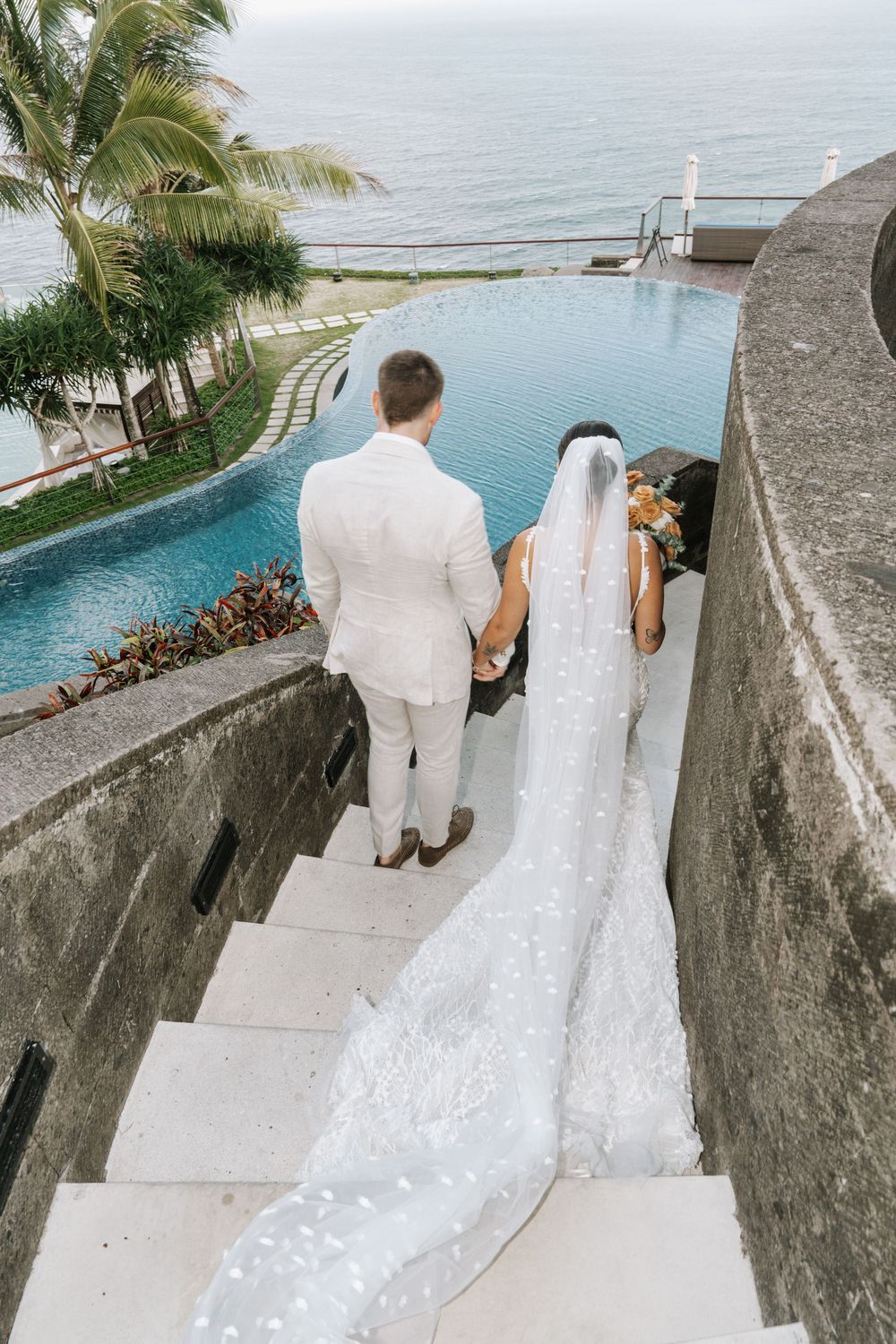 A couple descends stone steps beside an infinity pool overlooking the ocean with palm trees and tropical landscaping.