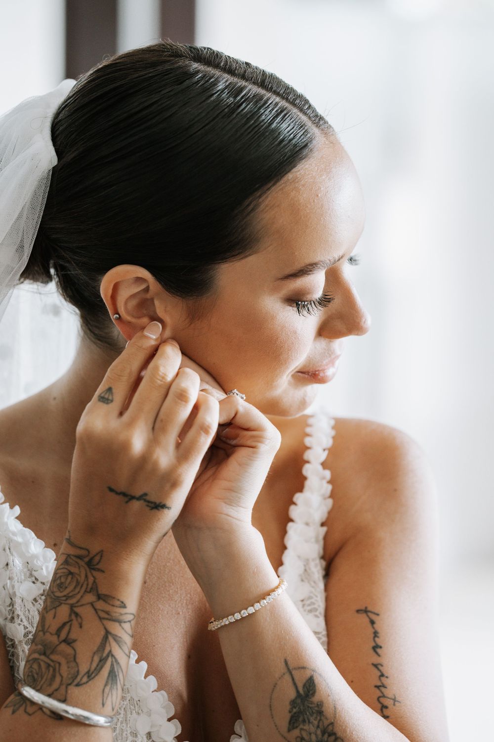 Side profile of a bride in white lace gown putting on earrings, showing delicate tattoo details in elegant bridal portrait.