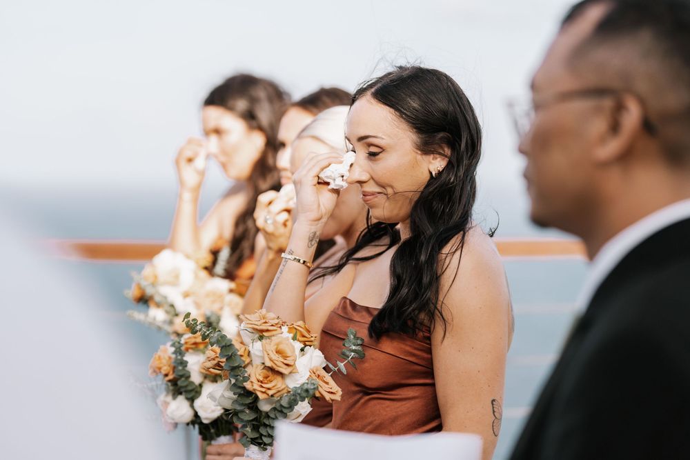 Wedding party attendants stand in a row near a waterfront venue in formal attire.