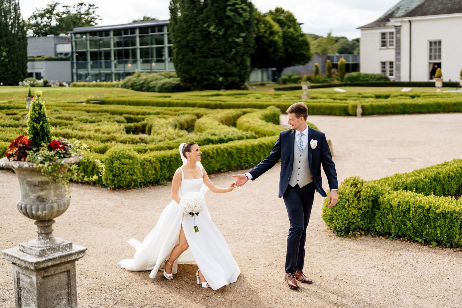 A wedding party walks through an elegant garden maze with perfectly manicured hedges and stone pathways.