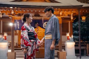 Two people in traditional Japanese kimonos stand in front of illuminated temple buildings at dusk.