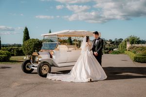 Wedding couple poses near a classic vintage car during golden hour photography session at a scenic outdoor venue.
