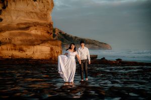 A couple in wedding attire stands on rocky coastal terrain at sunset with dramatic cliffs in the background.