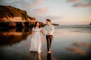 Two figures in elegant attire share a romantic moment by the water during sunset at a scenic beach location.