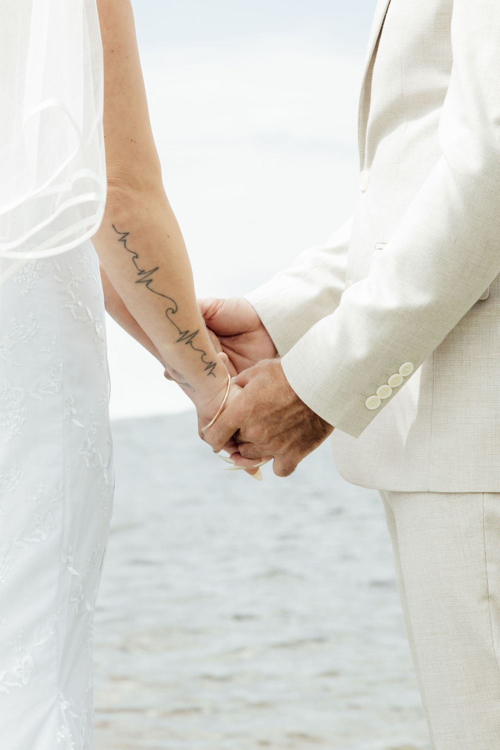 Two people in white attire holding hands with tattoos on their wrists at what appears to be a beach setting.