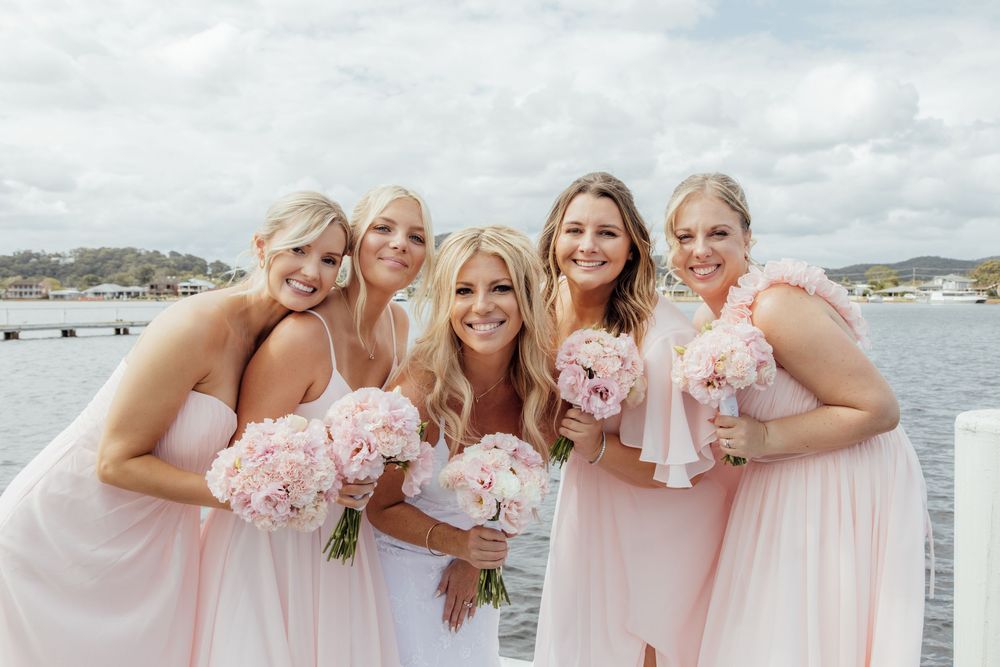 Bridesmaids in blush pink dresses holding light pink bouquets pose together by the waterfront on a cloudy day.