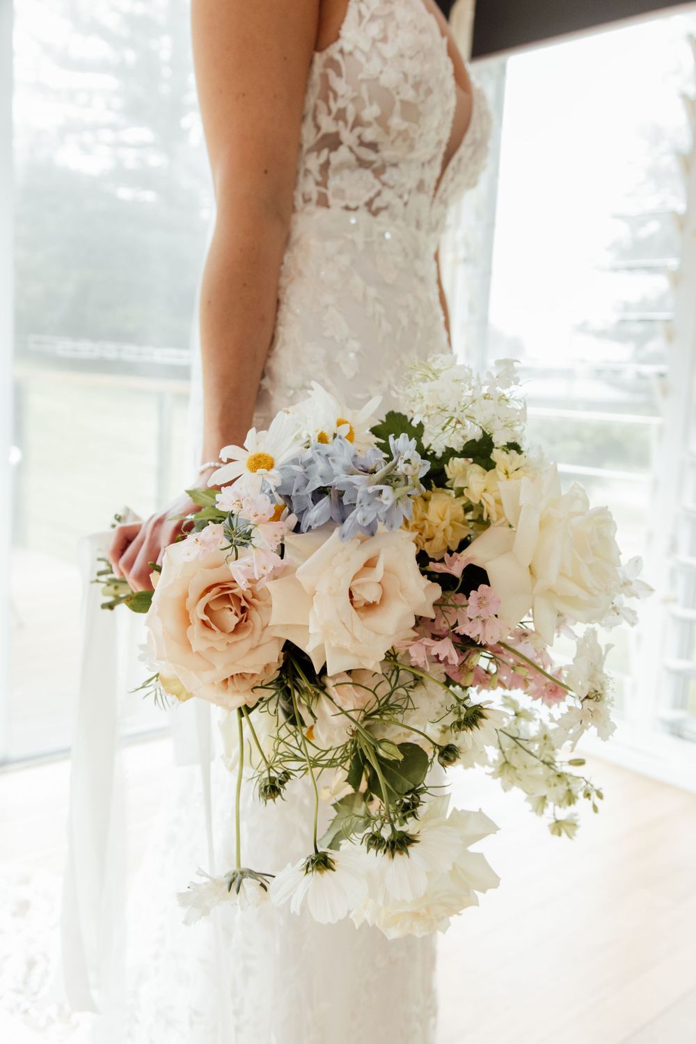 A romantic bridal bouquet with blush roses, white daisies, and delicate blue flowers against a lace wedding dress.