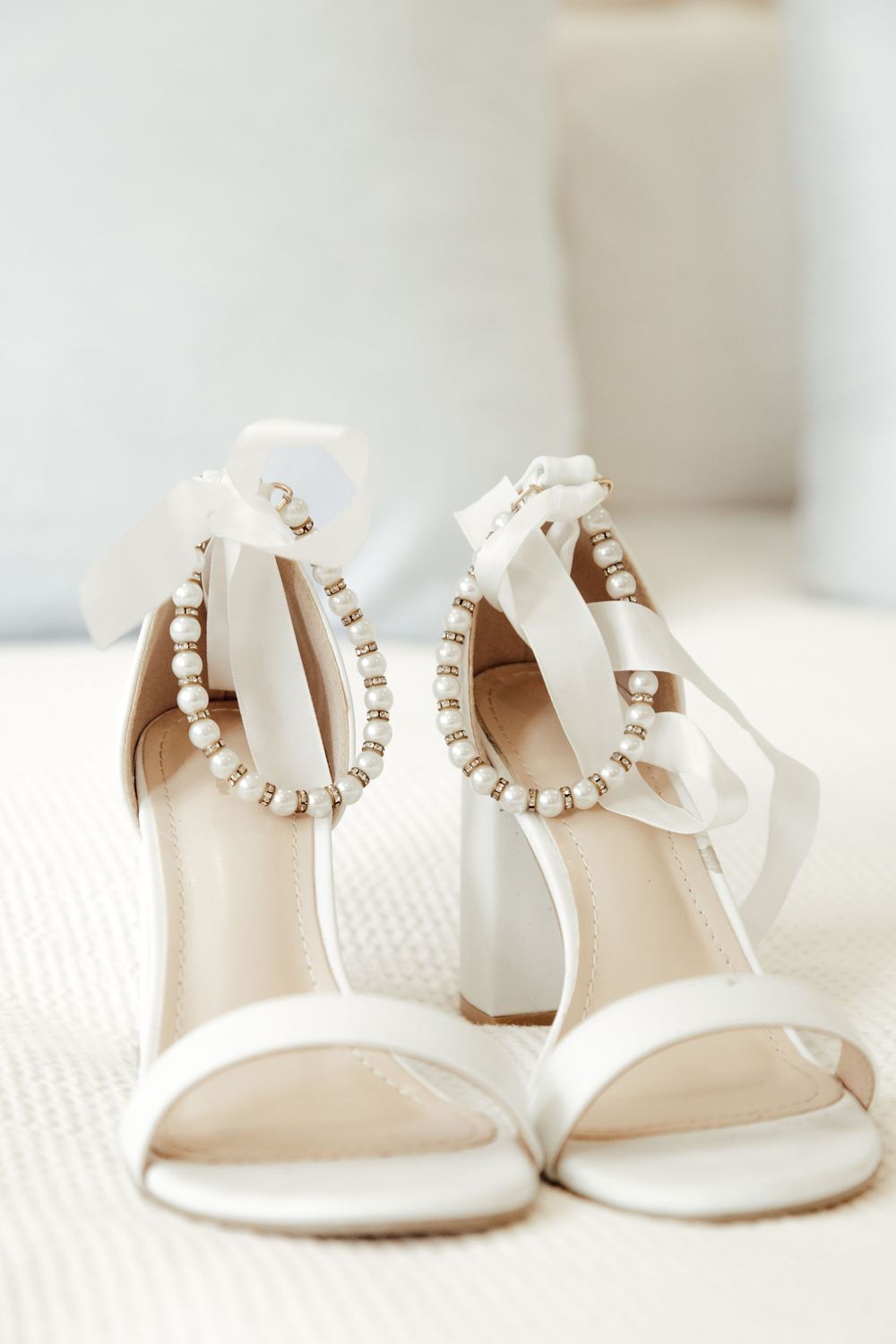 Elegant white wedding sandals with pearl ankle straps and ribbon details sit on a light textured surface.